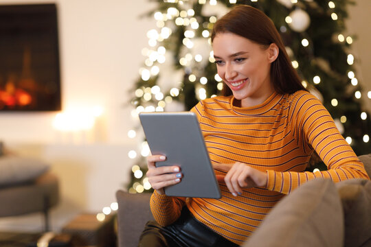 A cheerful young woman sits on a couch, engaged with her tablet. The room is warmly lit with holiday lights, creating a festive atmosphere.
