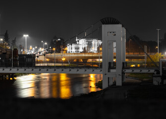 night view of a modern suspension bridge and illuminated government building over a river, with golden water reflections in the city.