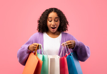 A joyful young black woman opens her shopping bags on a vibrant pink background. She enjoys the thrill of discovering items she purchased, celebrating great deals and discounts.