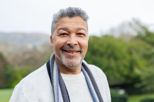 Mature adult man standing in park wearing white knit sweater and striped scarf under overcast sky