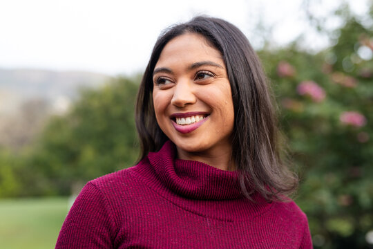 Asian woman standing in garden wearing maroon turtleneck and smiling at pink blossoms
