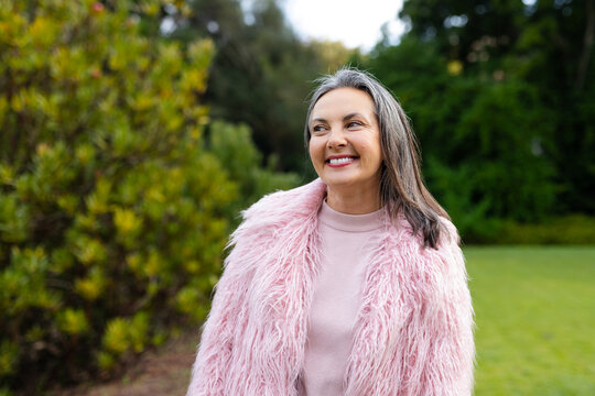 Woman standing wearing pink faux fur coat and smiling while gazing left in manicured garden