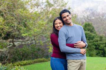 Diverse couple wearing knit sweaters hugging on green lawn in park-like garden under overcast sky