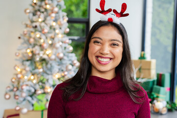 Asian woman smiling wearing antler headband standing in living room by frosted tree near gifts