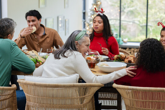Diverse family gathering in dining room at table sharing roasted turkey, salads, rolls and drinks