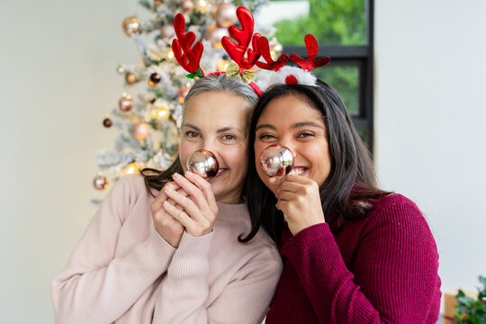 Diverse mother and daughter holding baubles near noses wearing antler headbands by Christmas tree