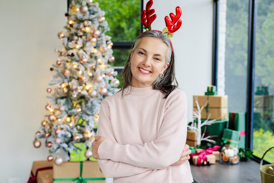 Woman wearing reindeer antler headband crossing arms, smiling by Christmas tree with gifts at home