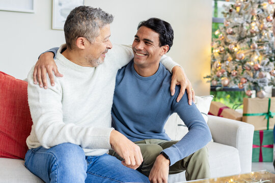 Father and son sitting on sofa in living room, smiling by Christmas tree with gifts