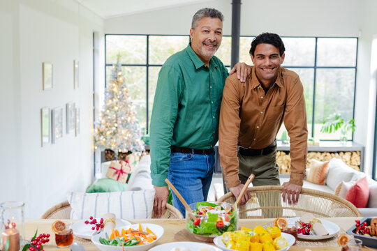 Father and son smiling behind dining table in living room with Christmas tree and wrapped gifts