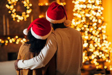 Obraz premium A loving young black couple hugs while wearing Santa hats and cozy sweaters. They gaze at a brightly decorated Christmas tree, enjoying their quiet holiday evening together.