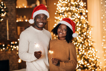 Romantic young black couple wearing comfy sweaters and Santa hats joyfully holds sparklers while...