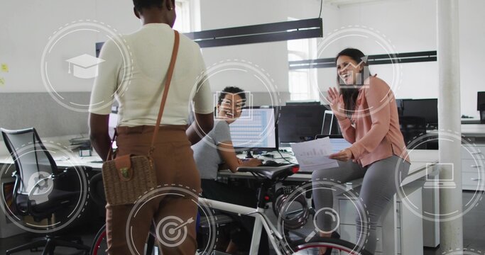 Three coworkers pedaling desk bike and waving papers in office near monitors, with floating icons
