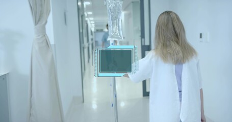 Walking patient with wristband in gown pushing IV drip stand down white corridor, sterile ward