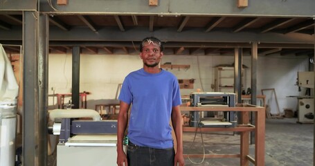 Standing carpenter wearing safety glasses holding tape measure in woodworking workshop with planer