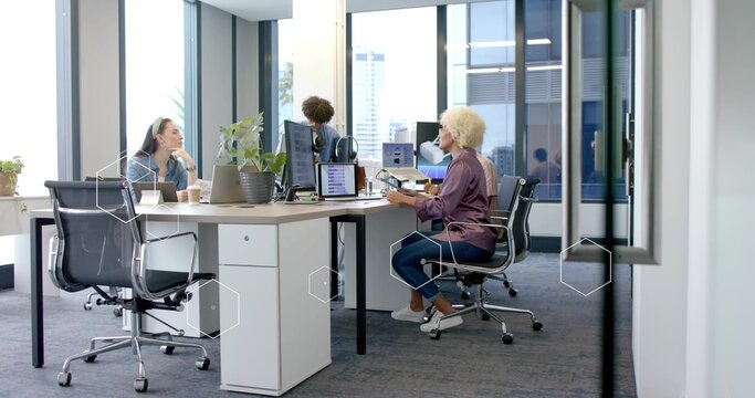 Collaborating coworkers typing on laptops at open-plan office desk, with coffee cups and monitors