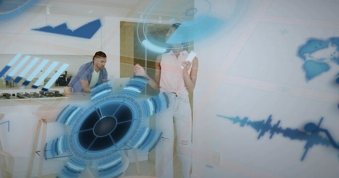 Woman holding cards and gesturing toward hologram at kitchen island, man leaning by laptop