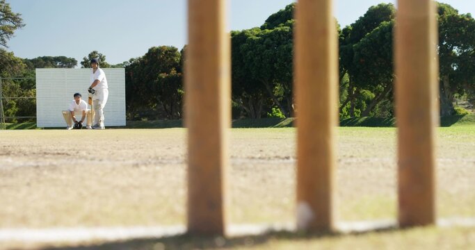 Framing wooden stumps occupying center at pitch, batter in pads helmet holding bat, keeper gloving
