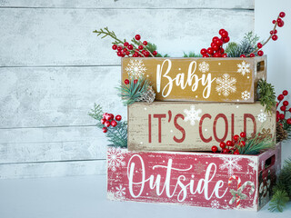Old farm Christmas country stacked crates with holiday text Baby It’s Cold Outside and festive evergreen greenery branches and red berries on rustic white light wood plank backdrop.