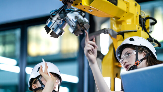 Two female engineers in hard hats and safety vests collaborate in a factory. One woman holds a laptop while another points at a large yellow industrial robotic arm.