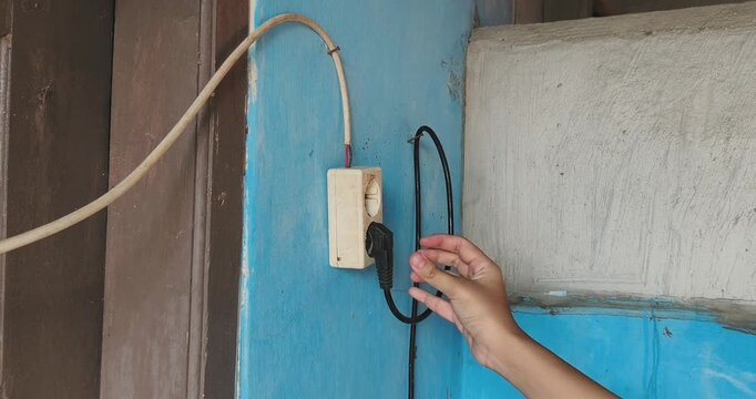 Hand plugging the black power cord into the old white wall socket on the bright blue wall. 