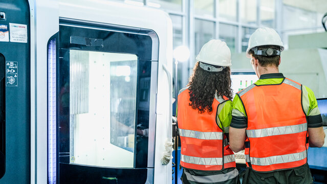 Two engineers use a laptop to program an industrial 5-axis CNC machine. They are programming an AI-driven system for high-precision component manufacturing.