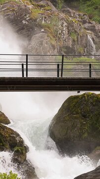 Woman hiker crosses a bridge over the Kleivafossen waterfall on the way to Brikdalsbreen Glacier in Norway. Vertical Video.