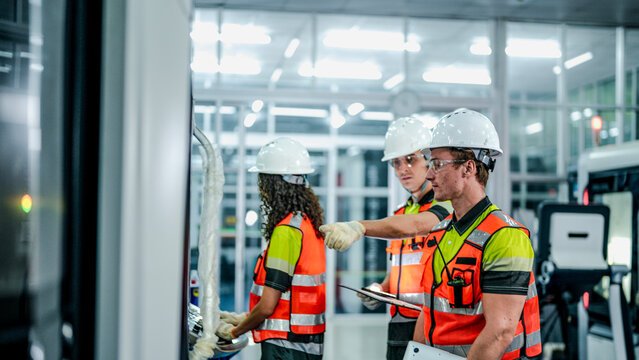 A team of engineers in hard hats and safety vests operates a high-tech industrial machine. The man in the foreground programs the advanced equipment in a smart factory.