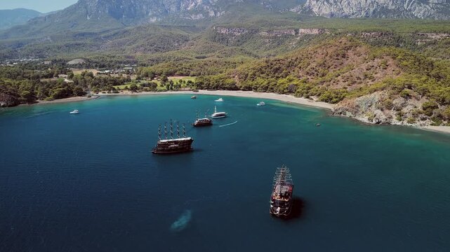 Aerial view of Phaselis Bay with sailing ships and Mount Olympos in the background. Clear blue water and scenic Mediterranean coastline near Antalya, Turkey. Captures scenic coastal beauty in Turkey.