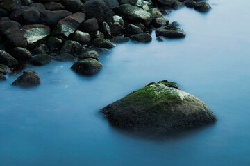 A large, moss-covered stone emerges from the smooth, misty blue water near a dark, rocky shore.