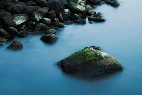 A large, moss-covered stone emerges from the smooth, misty blue water near a dark, rocky shore.