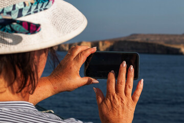 Fototapeta premium A woman wearing a straw hat uses a cell phone to photograph a distant island over the dark blue sea during a sunny vacation trip.