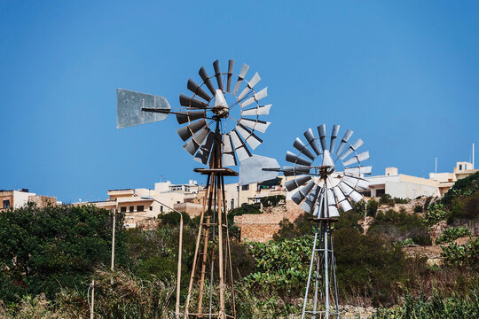Two multi-bladed metal windpumps stand prominently against a vivid blue sky, overlooking low-rise buildings and arid vegetation in a sunny climate.