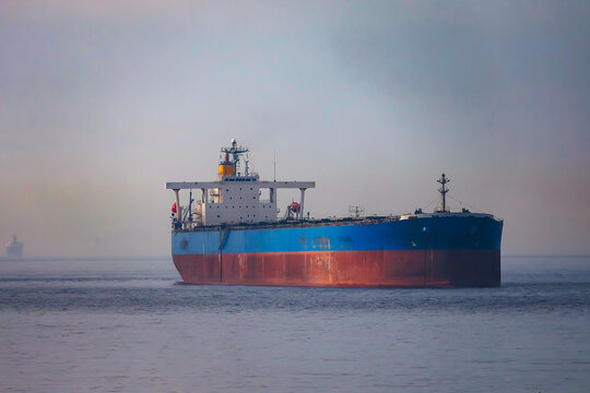 A large cargo ship, painted blue and red, sails through hazy calm seas under an overcast sky, with another vessel visible far in the distance. - Powered by Adobe