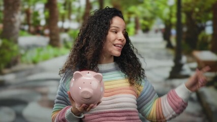 Woman smiling outdoors holding pink piggybank in a colorful sweater on a vibrant street indicating savings suggestion in a cheerful urban setting.