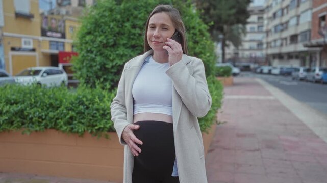 Pregnant woman talking on phone on urban street with trees and buildings in the background, embodying modern lifestyle and expecting motherhood outdoors.