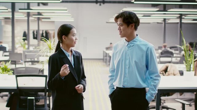Two young Asian professionals converse in a contemporary office environment filled with plants and natural light. Their attire suggests a formal work setting, indicative of a collaborative atmosphere.
