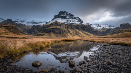 Snow capped mountain range with rocky cliffs reflecting in calm water pool surrounded by dry grassland under moody cloudy sky landscape