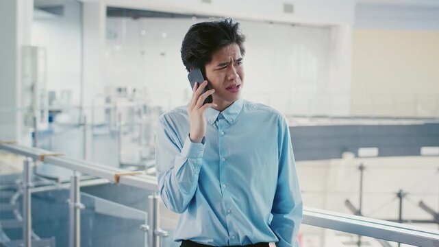 A young Asian man is engaged in a phone call, appearing concerned as he stands in a contemporary indoor location. The background features sleek architecture and bright lighting.