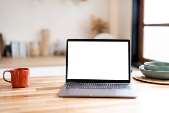 Laptop with a blank white screen placed on a wooden table next to a coffee mug and breakfast bowls in a home kitchen setting.