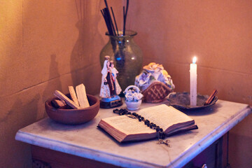 View of a small home altar with lit candles, a Bible, a cross, decorations, and a statue of the Virgin Mary, all bathed in warm light. From an olden time.