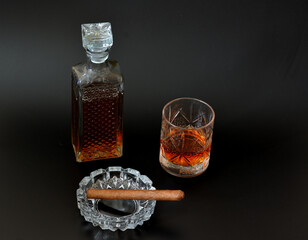 A faceted glass and a crystal decanter with old whiskey and a glass ashtray with a Cuban cigar on a black background.