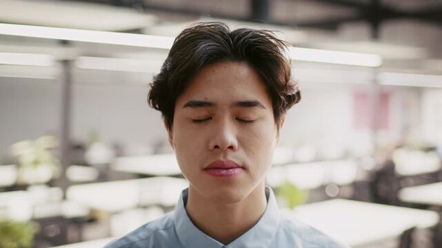 A young Asian man sits quietly in a contemporary office space. The minimalist design includes plants and ample natural light, enhancing a calm atmosphere for focus.