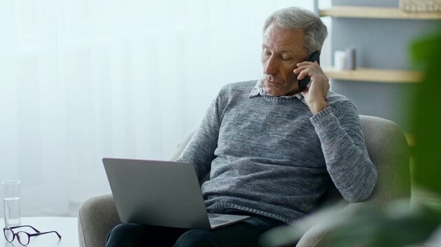 An elderly man in his 60s sits comfortably in an armchair. He speaks on the phone, while a laptop rests on his lap, reflecting a modern lifestyle focused on home and work.