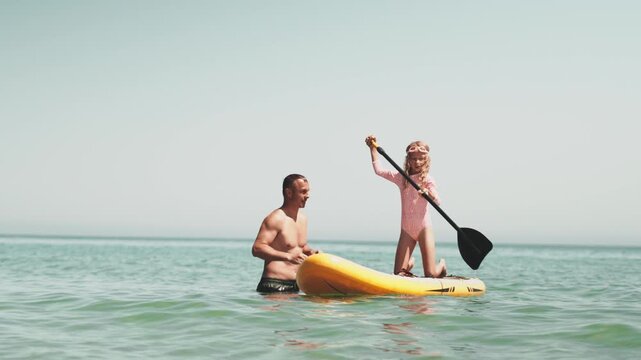 Dad and little girl paddle boarding on calm sea during family summer holiday, enjoying time together under bright sunlight. Dad teaches Daughter to swim on Sup