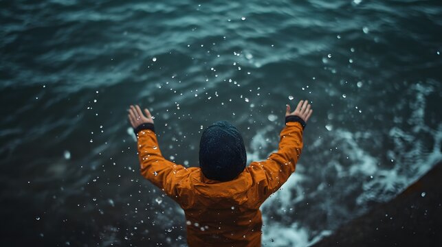 A person with arms outstretched stands on a rocky shore with waves crashing behind them.