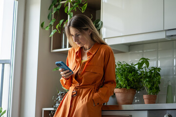 Relaxed woman holding phone, standing in cozy kitchen near window, leaning on counter with house...