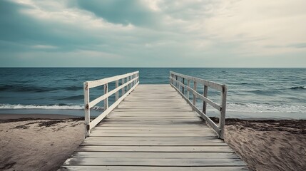 Fototapeta premium A wooden pier extends into the ocean, with a sandy beach and cloudy sky in the background.