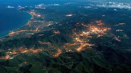 Coastal cityscape at night illuminated by streetlights with mountain ranges surrounding urban areas and glowing lights reflecting on calm ocean shore under partly cloudy sky.