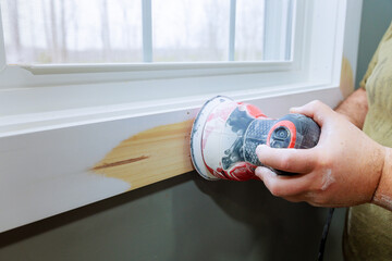 Carpenter is sanding wooden trim piece near window as part of room renovation dust wood grain.