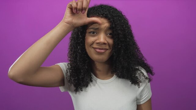 Woman lifts hand making l sign to forehead and frowns in purple studio showing curly hair; mockery teasing.